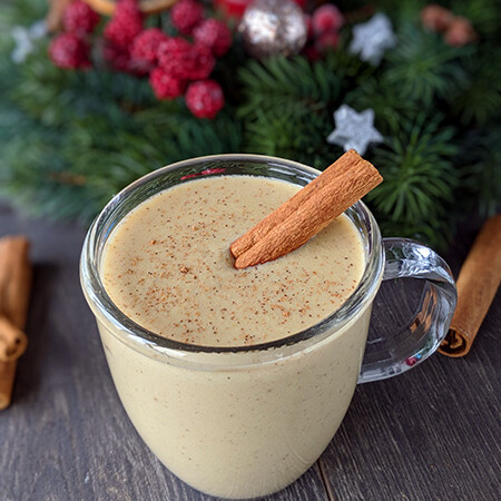 photo of a cup of creamy eggnog with a cinnamon stick and holiday decorations in the background