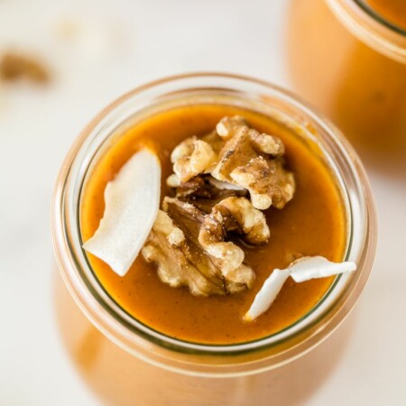 overhead photo of a jar of pumpkin pudding that is sweetened with honey garnished with coconut slivers and toasted walnuts