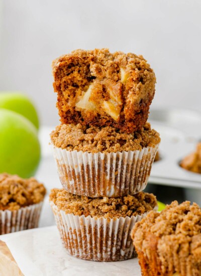 Photo of three healthy cinnamon apple muffins stacked upon each other with a bite taken out of the top muffin showing chunks of apples in the middle of the muffin