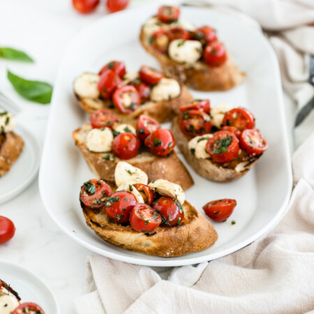 Photo of toasted bruschetta with mozzarella and halved cherry tomatoes with fresh basil and a little oil on a white platter