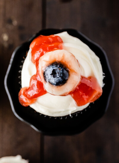 top view photo of Eyeball Cupcakes for Halloween that have a moist chocolate base, cream cheese frosting and eyeballs that consist of canned lychees, and blueberries and strawberry jam to look like “blood” dripping down the frosting.