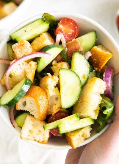 Photo showing a bowl full of green Tuscan-style panzanella salad loaded with chunks of bread, fresh ripe tomatoes, spicy arugula, crunchy cucumbers, sweet red onions, and zesty capers.