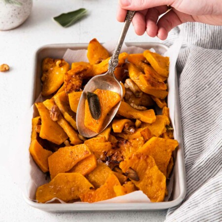 Image of a hand with a serving spoon dipping into a dish of creamy, buttery, nutty baked butternut squash seasoned with fresh sage, garlic cloves and crunchy walnuts