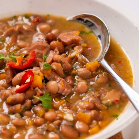 Image of a spoon in a bowl of hearty Instant Pot Bean Soup make with sautéd onions, carrots, diced ham with plenty of beans garnished with sliced red pepper and cilantro.