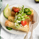 image of four crispy air fryer taquitos on a white plate garnished with lettuce and halved cherry tomatoes and two slices of jalapeños on a white plate with two forks and two slices of green avocado