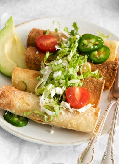 image of four crispy air fryer taquitos on a white plate garnished with lettuce and halved cherry tomatoes and two slices of jalapeños on a white plate with two forks and two slices of green avocado