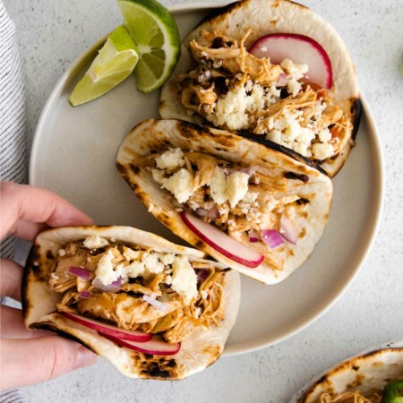 image showing a hand grabbing one of three instant pot shredded chicken tacos garnished with slices of radish and a bit of cheese with two lime slices on the side of the white plate