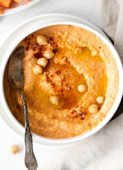 close up image of roasted red pepper hummus garnished with a few whole chickpeas and a dusting of smoked paprika on top with a spoon set in the bowl