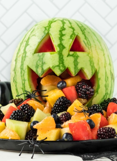 image of a watermelon cut Jack-o-lantern style with fresh fruit and berries spilling out of the mouth as a fun Halloween fruit salad with a few fake spiders crawling on the fruit