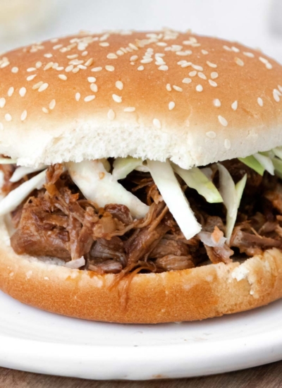 image of an instant pot pork shoulder sandwich showing perfectly seasoned pork shoulder on a hamburger bun garnished with raw cut green cabbage on a white plate