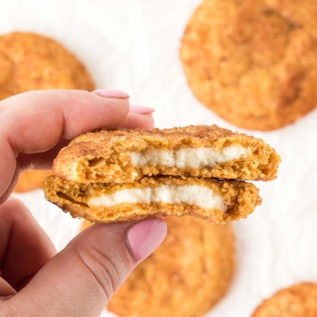 hand holding a pumpkin cheesecake cookie with a bite out of it showing the layers of cookie around cheesecake filling with more cookies on parchment paper in background