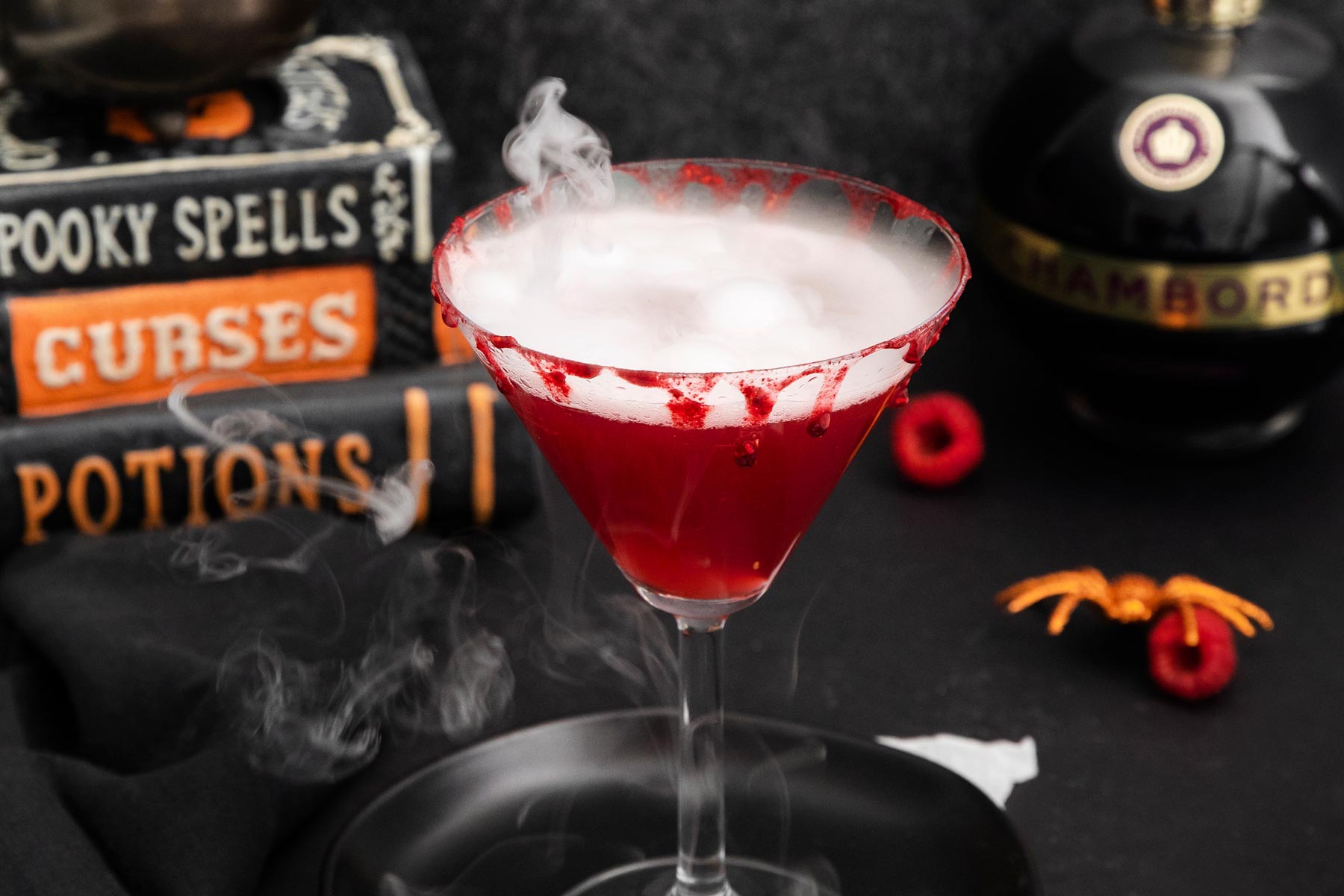 A spooky red cocktail in a martini glass with a smoke effect, against a dark backdrop. The glass rim is decorated with red sugar. In the background, there are Halloween-themed items including books labeled "Spooky Spells" and "Curses," and a bottle of Chambord.