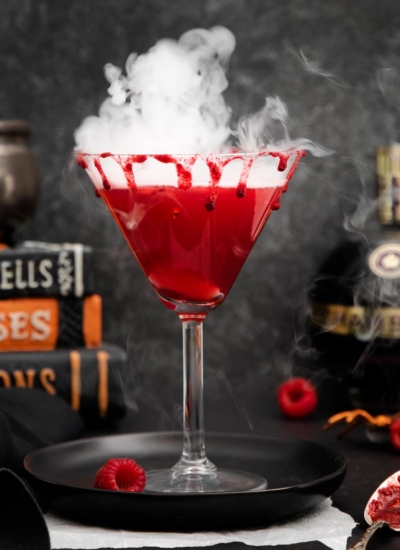 image of a martini glass filled with a Vampire's kiss cocktail that gets its gorgeous color from raspberries and the Chambord, smoking from dry ice and sitting on a black plate with raspberries scattered about with a bottle of Chambord and a stack of scary books in background