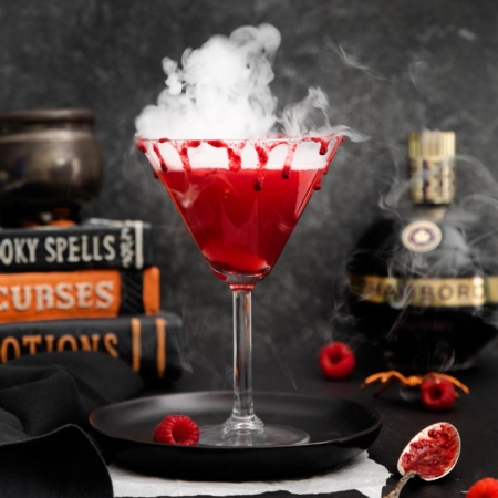 image of a martini glass filled with a Vampire's kiss cocktail that gets its gorgeous color from raspberries and the Chambord, smoking from dry ice and sitting on a black plate with raspberries scattered about with a bottle of Chambord and a stack of scary books in background