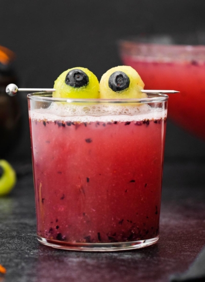 a glass of red non-alcoholic Halloween punch with green and blue eyeballs made from melon balls and blueberries with scattered melon balls in the background with a punch bowl full of punch