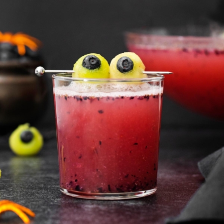 a glass of red non-alcoholic Halloween punch with green and blue eyeballs made from melon balls and blueberries with scattered melon balls in the background with a punch bowl full of punch