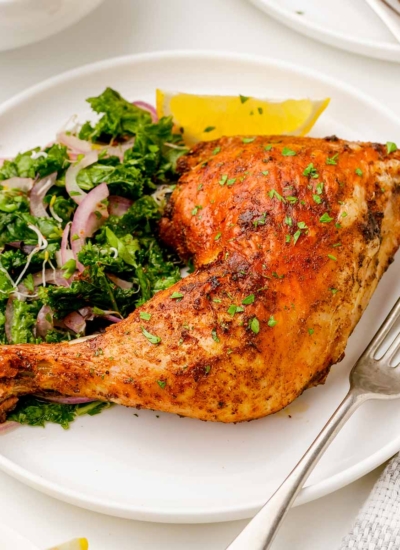 image of plated golden air fryer chicken quarter with a kale salad, lemon wedge and a fork on a white plate