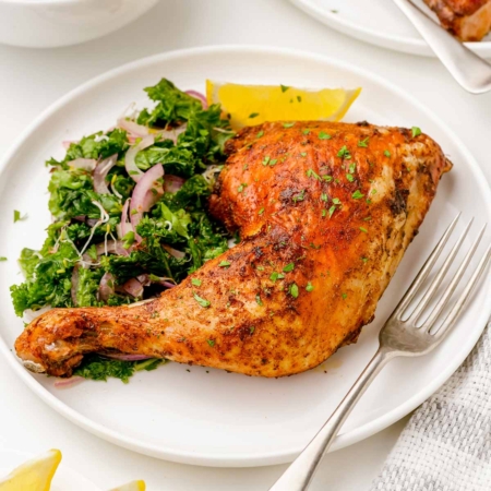 image of plated golden air fryer chicken quarter with a kale salad, lemon wedge and a fork on a white plate