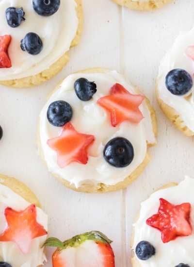 image of Memorial Day cookies using basic sugar cookie ingredients topped with a cream cheese frosting and strawberry star cut-outs with fresh blueberries giving it a red, white and blue theme scattered on a white background.