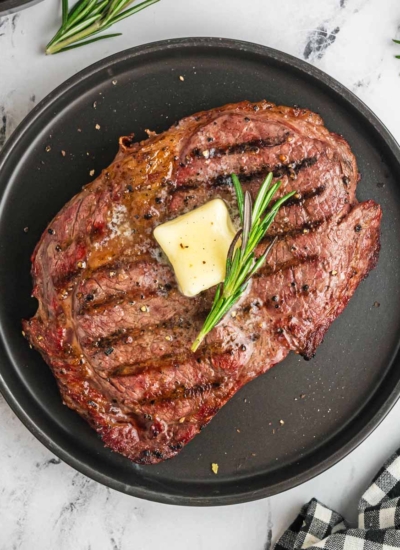 top view of a nicely seared smoked ribeye steak with perfect grill marks and a pat of butter on top with a rosemary sprig on a black plate and and white and black checkered cloth napkin to the side