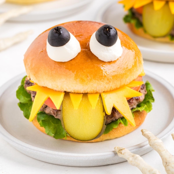 image of three adorable-looking monster burgers for Halloween, each assembled to look like a monster with lettuce, tomatoes on the bottom bun, and a juicy hamburger patty. It looks like a monster with sharp teeth made of cheese, a long sliced green pickle tongue hanging out from under the top bun, and two eyes made of mozzarella balls and black olives secured by a toothpick on top of the brioche hamburger bun to make the burgers look like cute monsters.