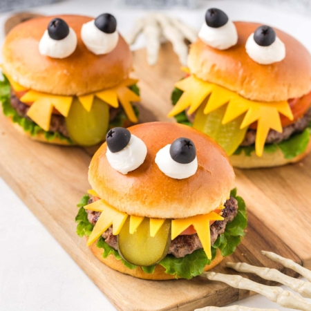 image of three adorable-looking monster burgers for Halloween, each assembled to look like a monster with lettuce, tomatoes on the bottom bun, and a juicy hamburger patty. It looks like a monster with sharp teeth made of cheese, a long sliced green pickle tongue hanging out from under the top bun, and two eyes made of mozzarella balls and black olives secured by a toothpick on top of the brioche hamburger bun to make the burgers look like cute monsters.