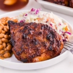 barbequed chicken thighs on a white plate with baked beans and coleslaw and a fork on the plate