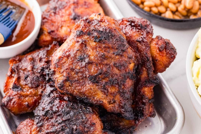 barbequed chicken thighs piled up on a baking pan with BBQ sauce in background