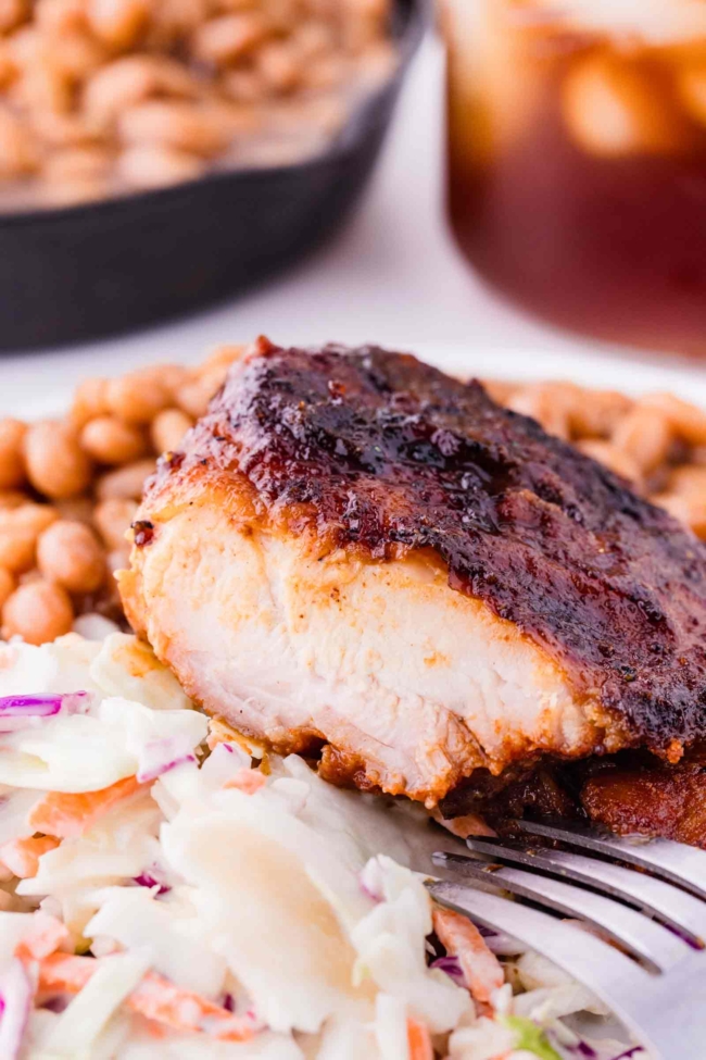 barbequed chicken thighs, cut into, on a white plate with baked beans and coleslaw