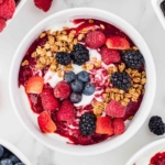 overhead shot of yogurt bowls with berries and granola on top with more berries in background