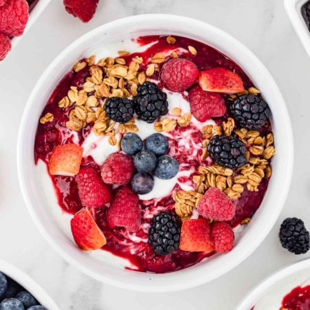 overhead shot of yogurt bowls with berries and granola on top with more berries in background