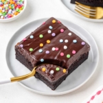 fork digging into a slice of a brownie with ganache on top on a white plate