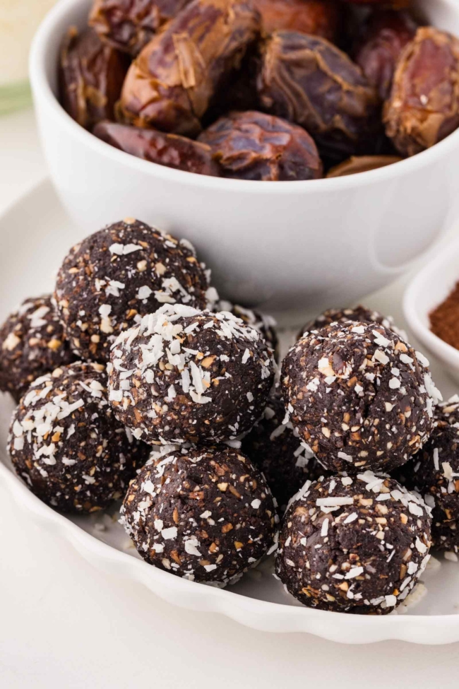 chocolate coconut nut balls on a white plate with bowl of dates in the background