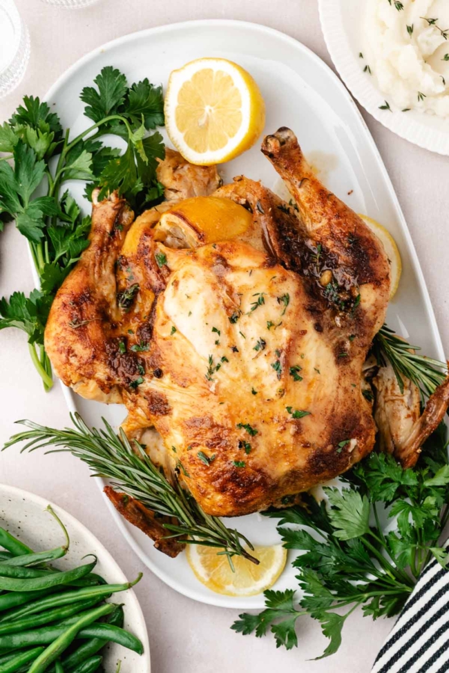 oven roasted whole chicken on a white serving dish with herbs surrounding it