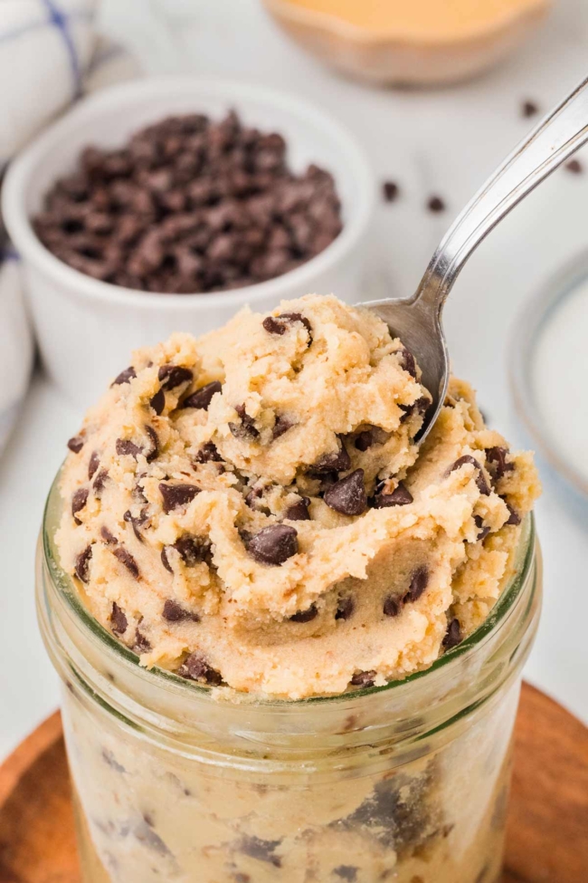 spoon digging into jar with cottage cheese cookie dough on a white table with bowl of chocolate chips in background