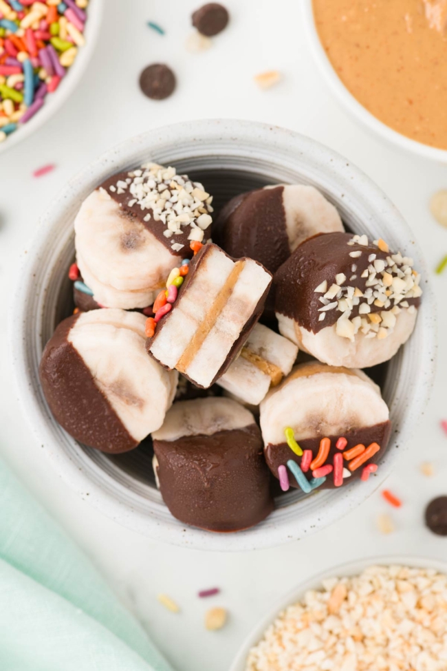 white bowl filled with peanut butter bananas on a white table with sprinkles and peanut butter in the background