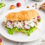 grape chicken salad in a croissant on a white plate with little tomatoes in background and pecan