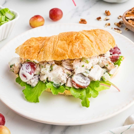 grape chicken salad in a croissant on a white plate with little tomatoes in background and pecan