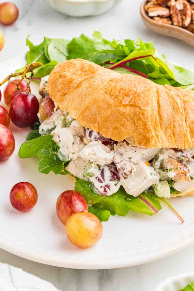 chicken salad in a croissant with lettuce on a white plate with tomatoes in background
