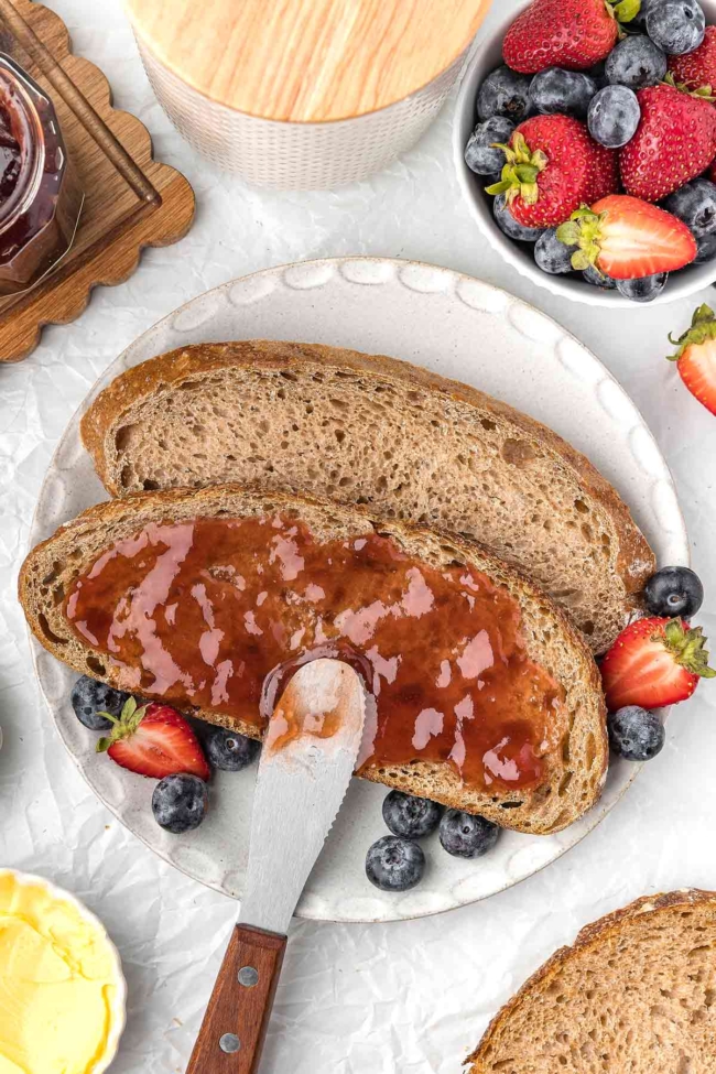 Whole wheat sourdough bread topped with jam, surrounded by fresh strawberries and blueberries on a plate.