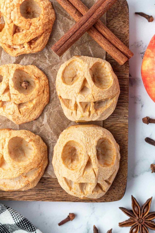 Carved apple halves baked into wrinkled shrunken heads, surrounded by cinnamon sticks and spices on a wooden board.