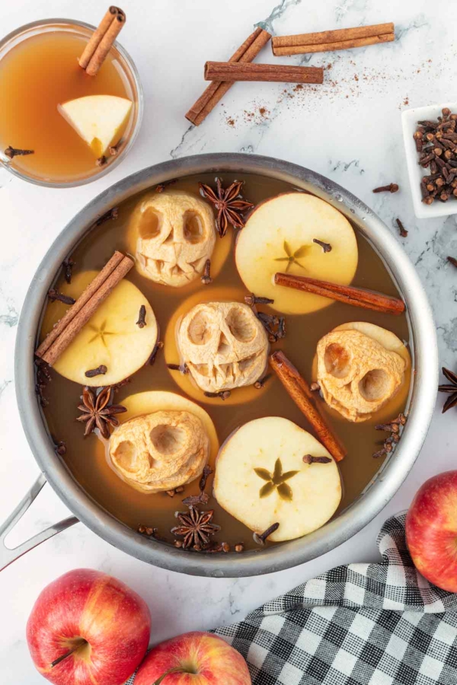 Spiced apple cider with carved apple shrunken heads, cinnamon sticks and star anise, surrounded by fresh apples and a checkered cloth.