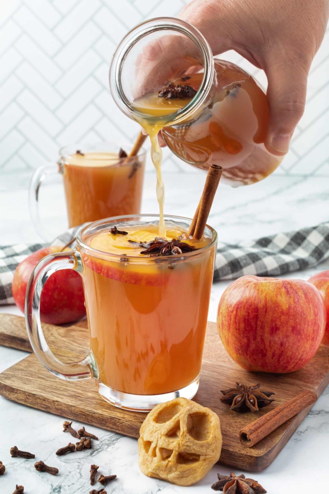 Warm spiced apple cider being poured into a glass with cinnamon sticks, star anise and fresh apples nearby.