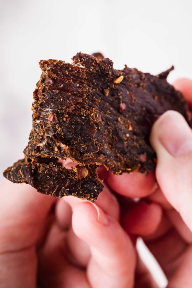 A hand bending a strip of venison jerky to demonstrate the cracks and separating meat fibers used to check dryness.
