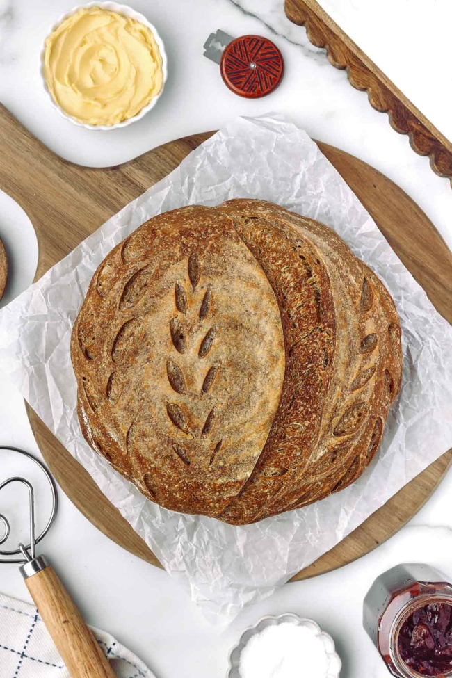 Whole wheat sourdough bread on parchment paper, surrounded by butter, jam, and baking tools on a wooden board.