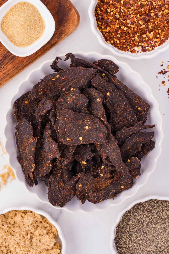 Savory venison jerky surrounded by various spices, including garlic powder, red pepper flakes, and black pepper.