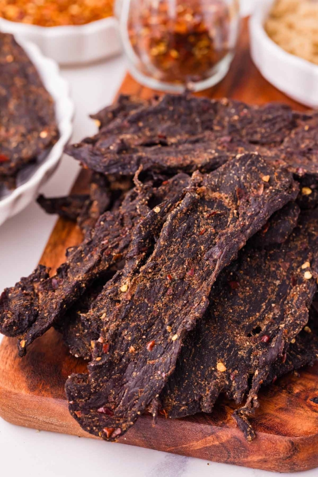 Savory venison jerky strips seasoned with spices, displayed on a wooden board with additional seasoning in the background.