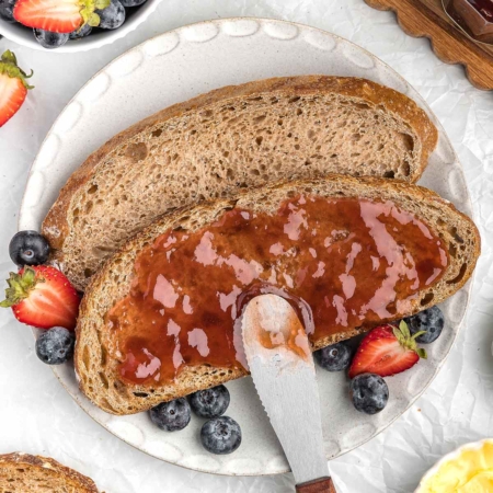 Whole wheat sourdough bread slice topped with jam, surrounded by fresh strawberries and blueberries on a plate.