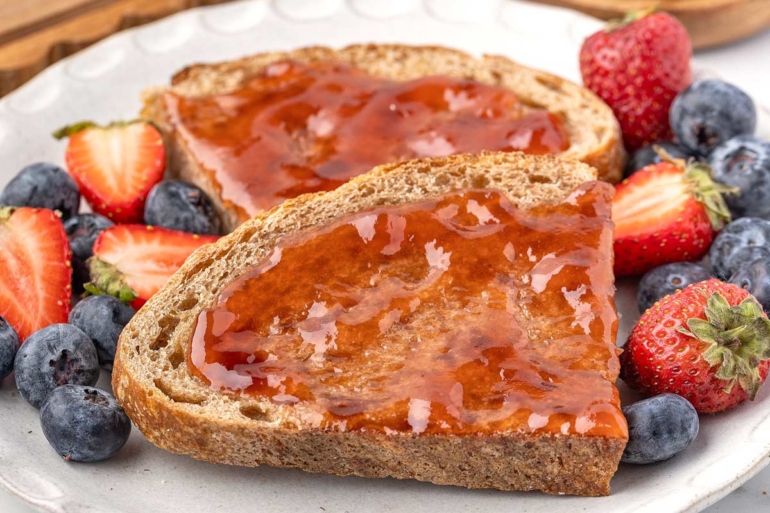 Whole wheat sourdough toast topped with jam, surrounded by fresh strawberries and blueberries on a plate.