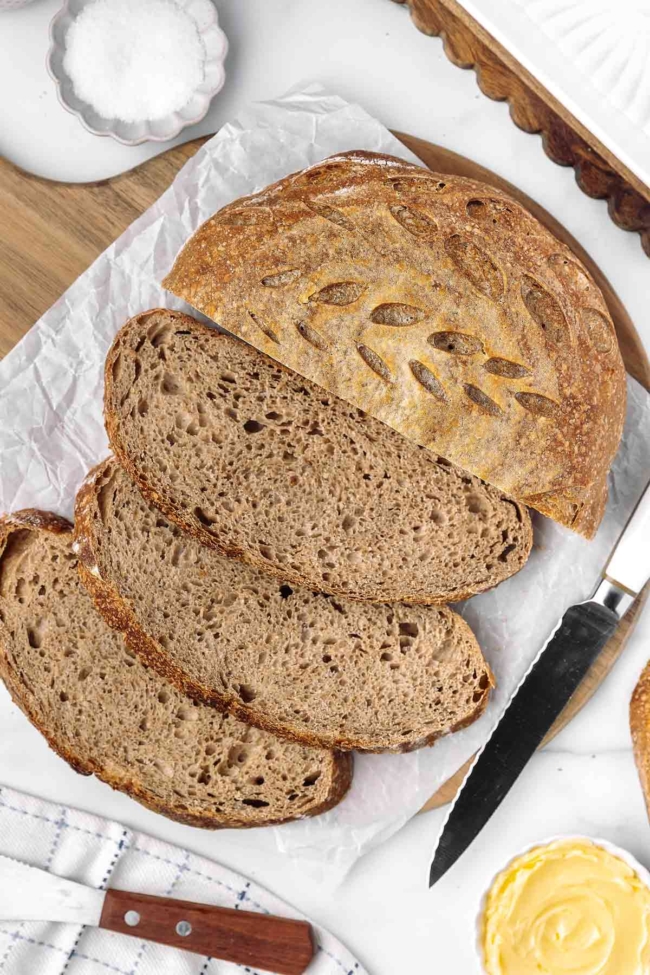 Whole wheat sourdough bread loaves and slices displayed on a wooden board with a knife and butter.